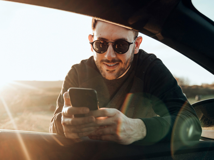 Ung man med solglasögon hänger in genom rutan till förarsätet i en bil, med sin mobiltelefon i händerna