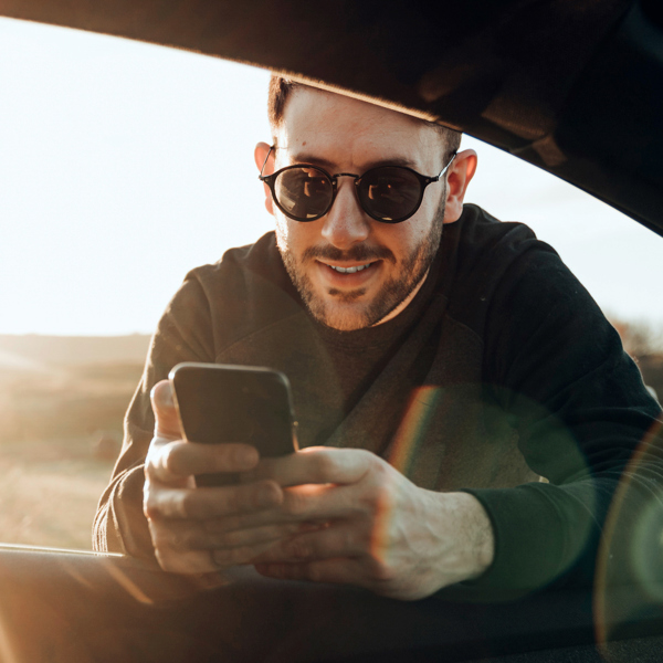 Ung man med solglasögon hänger in genom rutan till förarsätet i en bil, med sin mobiltelefon i händerna