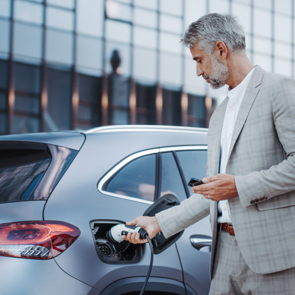 Gråhårig man med rutig grå kostym laddar sin elbil med ena handen medan han håller en mobil i den andra 